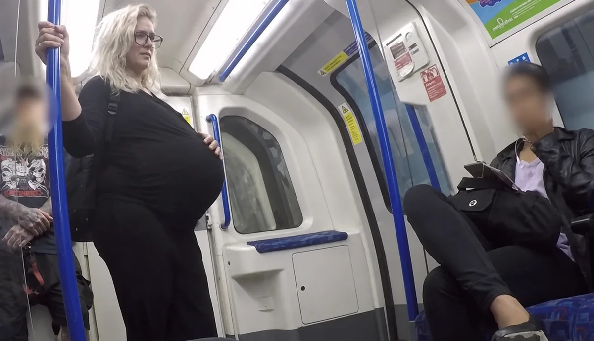 A heavily pregnant passenger standing and holding the pole on a tube train while the seated passengers either side look away
